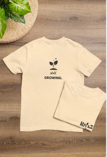Beige t-shirt with 'still growing.' text and logo on a wooden surface