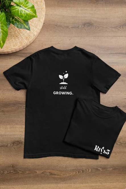 Black t-shirt with 'still growing.' text and plant graphic on a wooden surface.