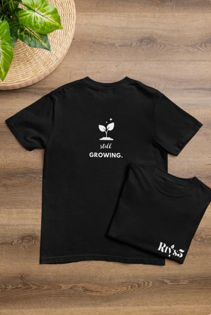 Black t-shirt with 'still growing.' text and plant graphic on a wooden surface.