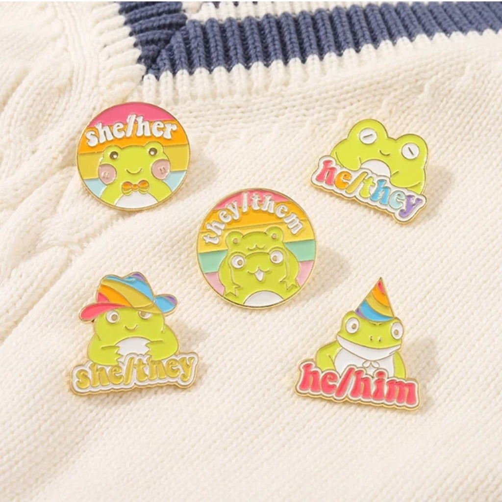 A collection of colorful enamel pins with cartoonish frog designs, showing pronouns like he/they, they/them, she/her, etc.